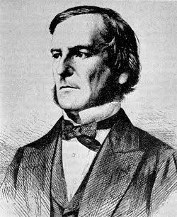 Homem branco, com longos cabelos grisalhos. De olhos claros e expressão serena. Veste terno escuro de estilo clássico.