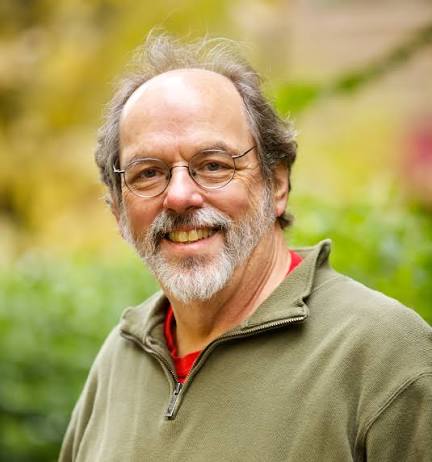 Homem branco de barba e cabelos grisalhos. Usa óculos redondos e veste uma camisa polo na cor cáqui. Apresenta-se com sorriso amistoso em um ambiente externo ensolarado.