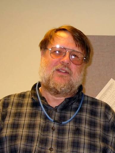 Homem idoso, branco, sorridente e de cabelos grisalhos. Usa barba e bigode brancos e óculos de grau com armação fina. Veste uma camisa quadriculada em tom marrom.