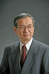 Homem de origem japonesa, pele clara, cabelos pretos curtos e grisalhos penteados para trás. Ele usa óculos de grau com armação retangular discreta. Veste terno cinza, camisa branca e gravata vermelha, apresentando uma expressão facial serena e sóbria.
