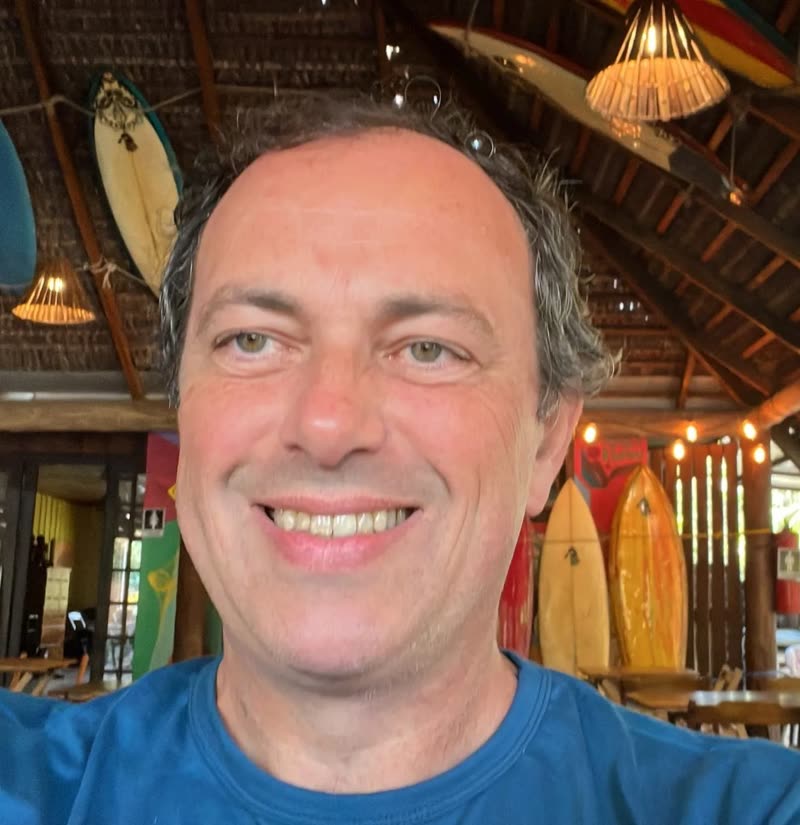 Homem branco de cabelos grisalhos e sorriso largo. Veste camiseta azul em ambiente descontraído, com pranchas de surfe coloridas ao fundo.