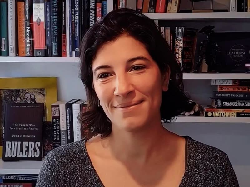 Mulher branca, com cabelos castanhos-escuros ondulados na altura dos ombros, leve sorriso, olhos escuros e blusa preta de gola canoa, com uma estante de livros ao fundo.
